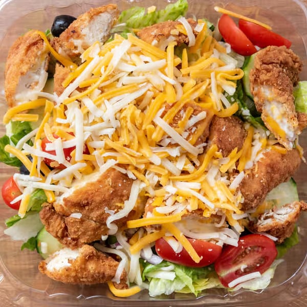 Crispy chicken salad with shredded cheese, lettuce, tomatoes, and black olives in a clear container.