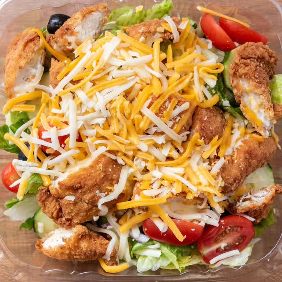 Crispy chicken salad with shredded cheese, lettuce, tomatoes, and black olives in a clear container.