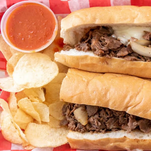 Two Giovanni's Pizza cheesesteak subs with chips and a side of marinara sauce on a red and white checkered paper.