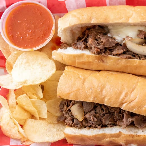 Two Giovanni's Pizza cheesesteak sandwiches with chips and a side of marinara sauce.